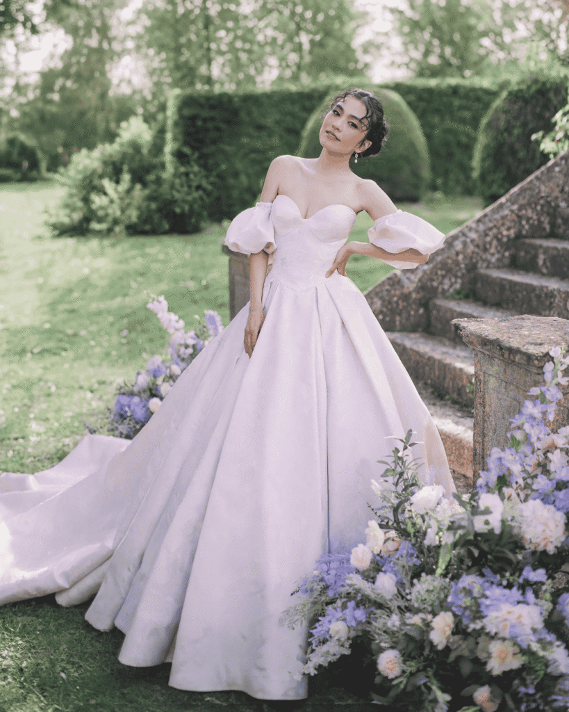 A pink wedding dress inspired by Edwina Sharma from Bridgerton, by Allure Bridals. The dress features a full skirt,sweetheart neckline, detachable sleeves, and subtle detailing on the fabric's surface.