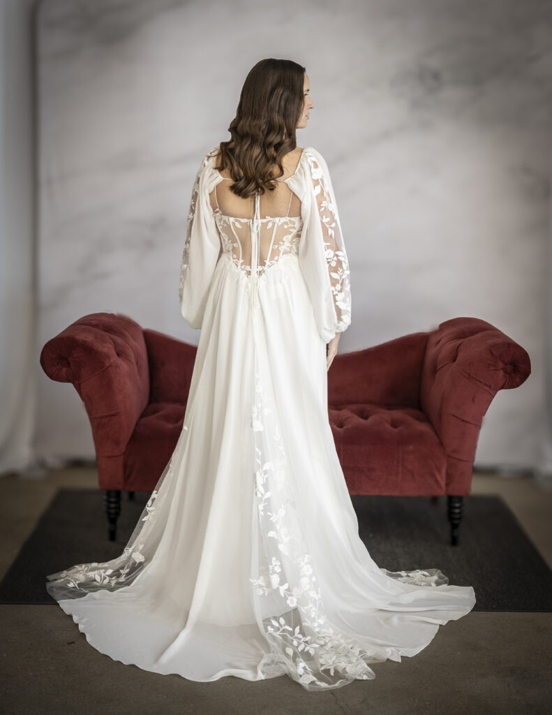 Rebecca Ingram's Sue wedding dress from the back perspective, showing off the corset-like bodice, the detachable sleeves, and the floral lace appliques down the sleeves and panels of the skirt.