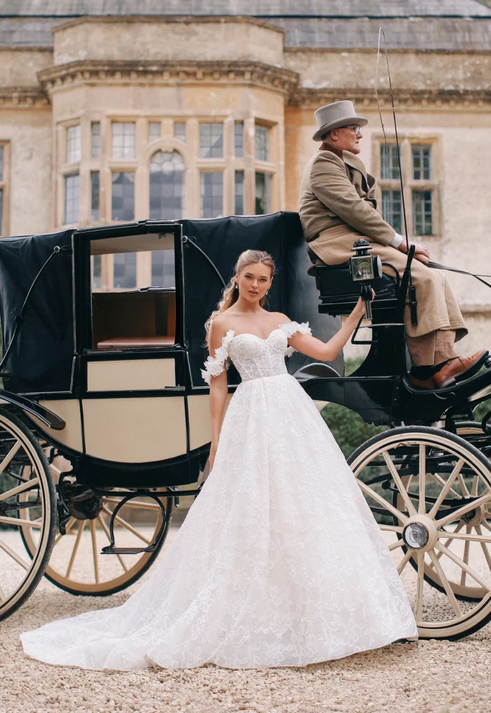 A model wearing the elegant and detailed Hyacinth wedding dress from the Allure Bridals Bridgerton collection, featuring frilly off the shoulder sleeves, an illusion lace bodice, and a full skirt.
